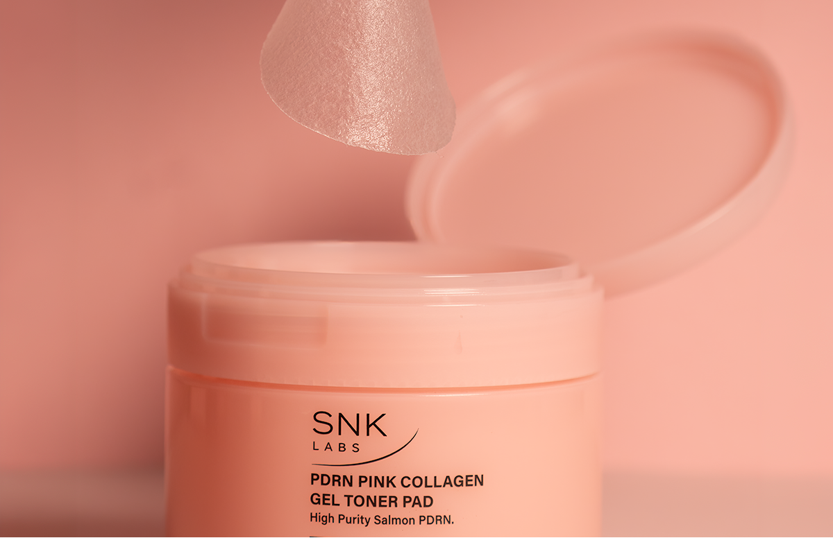 A close-up of a pink cosmetic jar with its lid open, and a translucent pink collagen toner pad being held above it by a hand. The jar label reads SNK LABS PDRN PINK COLLAGEN GEL TONER PAD.